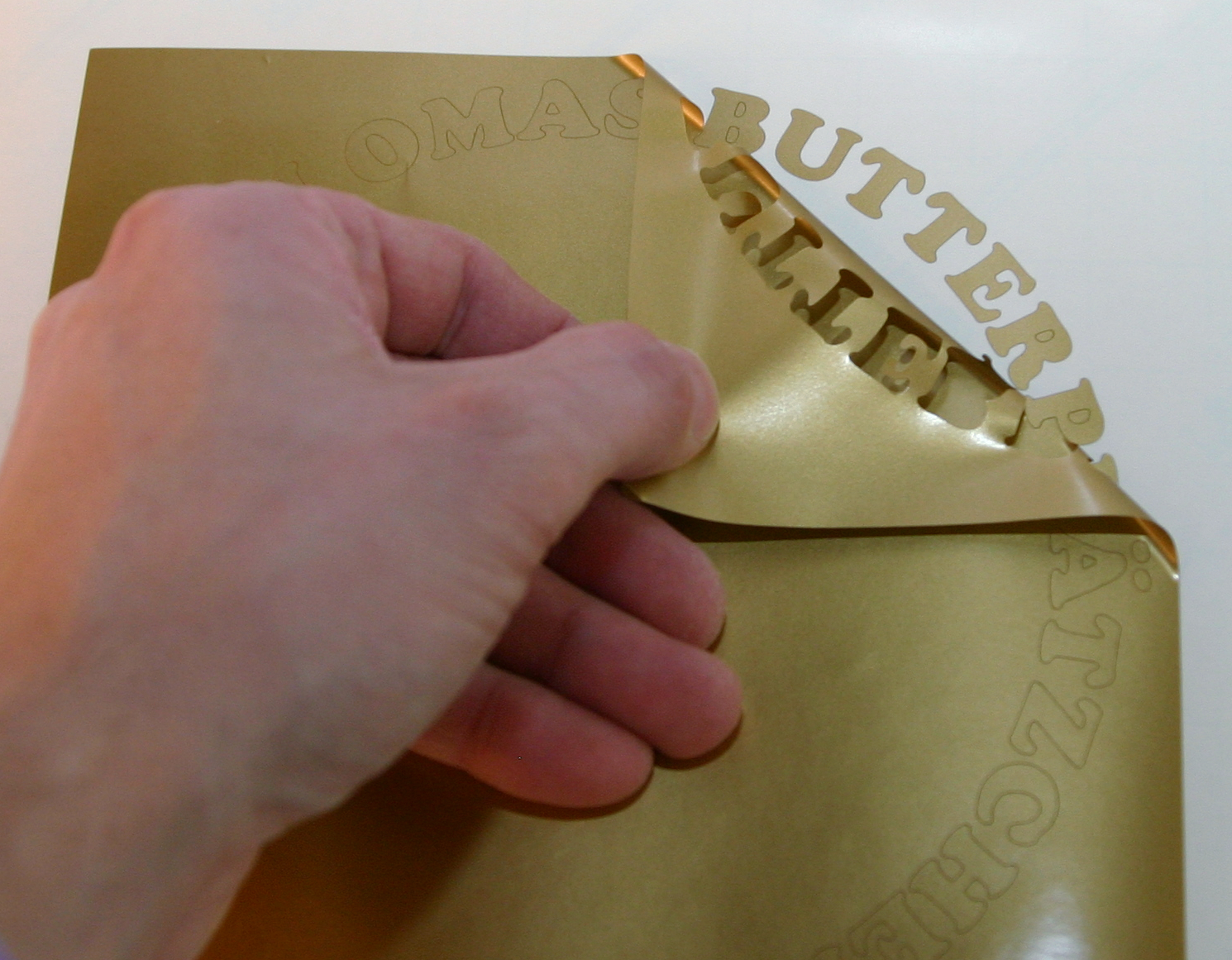 Hand zieht goldene Plotterfolie ab, der Schriftzug Omas Butterplätzchen bleibt im Bogen auf dem Trägerpapier: Entgittern des Aufklebers