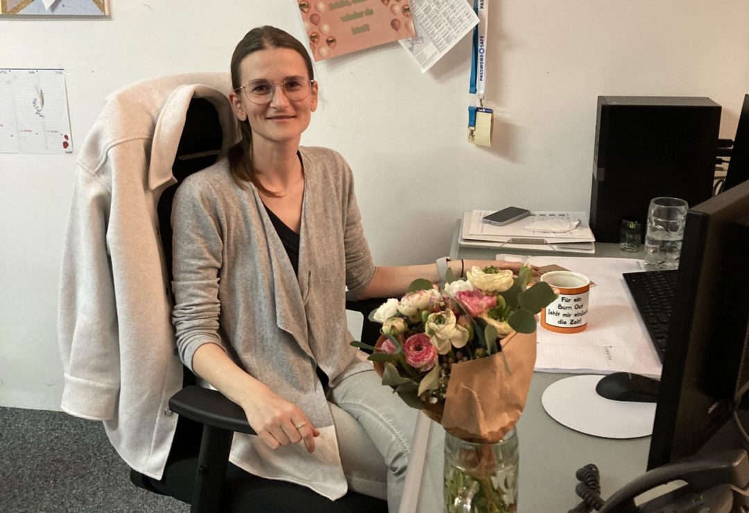 Moni lächelt an ihrem Schreibtisch im Büro, vor ihr ein Blumenstrauß zur Begrüßung nach ihrer Rückkehr