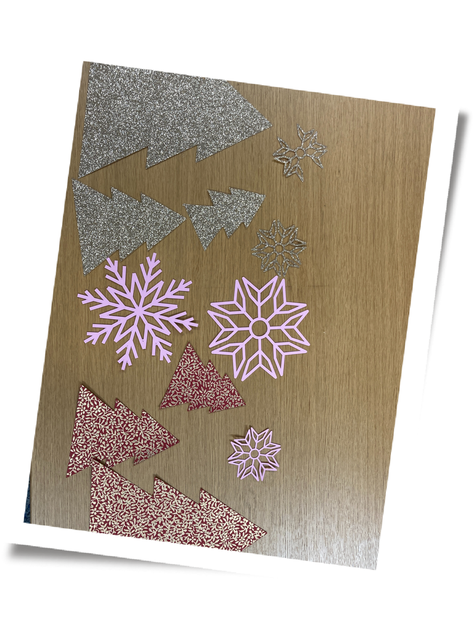 Sauber ausgeschnittene Tannenbäume aus Silberglitzer und rot-goldenem Papier sowie rosa und silberne Schneeflocken auf Holztisch