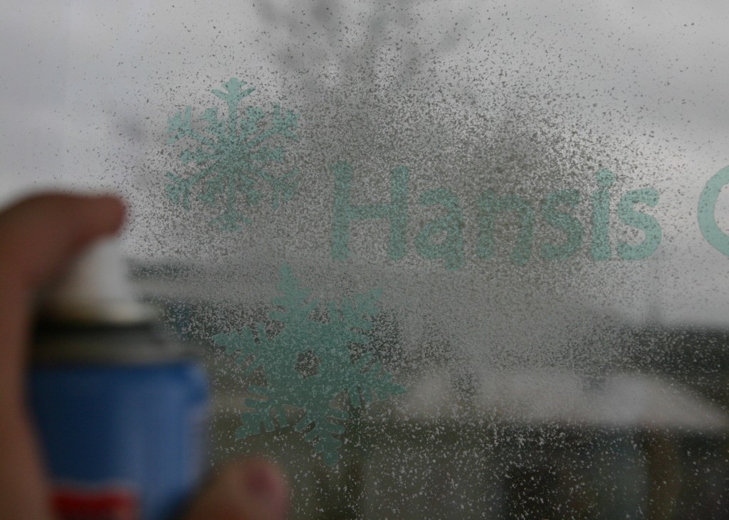 Hand besprüht Fensterscheibe mit Snow-Spray über aufgeklebte Maskierfolie mit Schneeflocken und Schriftzug Hansis