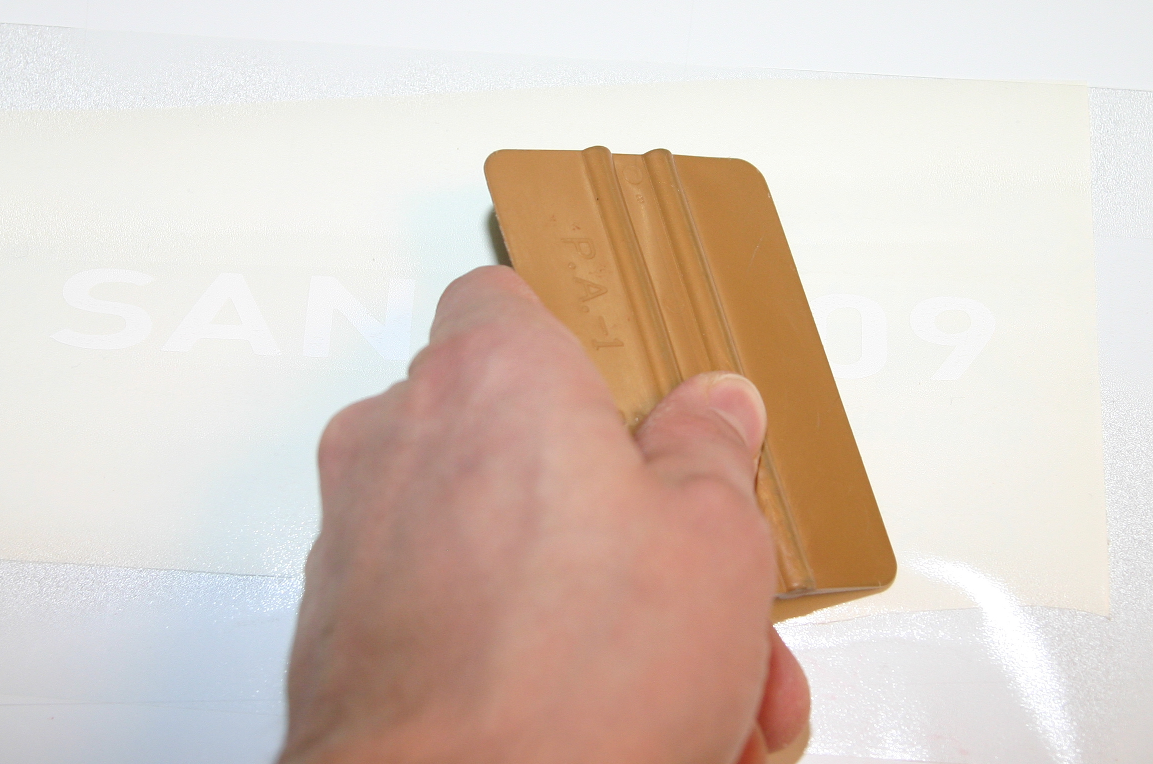 Hand rakelt mit goldenem Rakel die Übertragungsfolie mit weißem Plotterschriftzug SAN…09 fest, bevor die Folie abgezogen wird