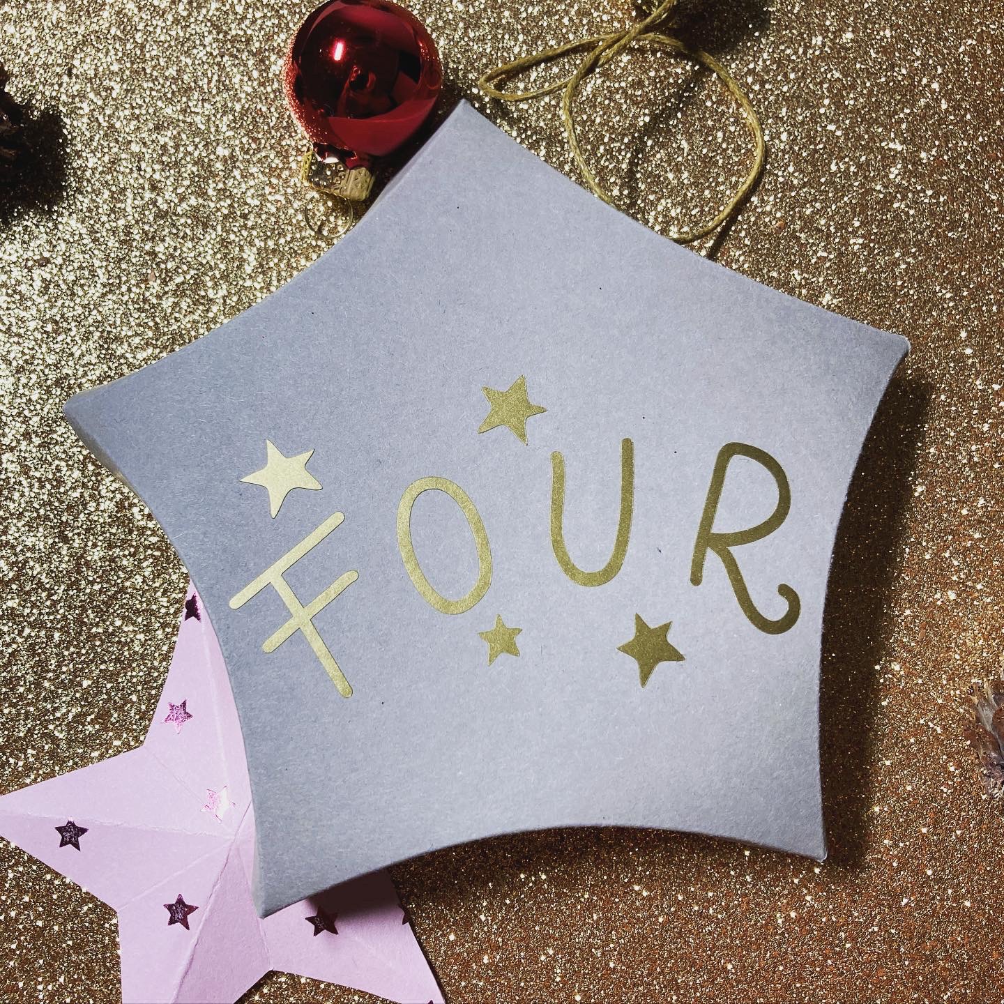 Sternförmige graue Schachtel mit goldener Aufschrift FOUR, daneben gefalzter rosa Papierstern auf Goldglitzer mit roter Christbaumkugel