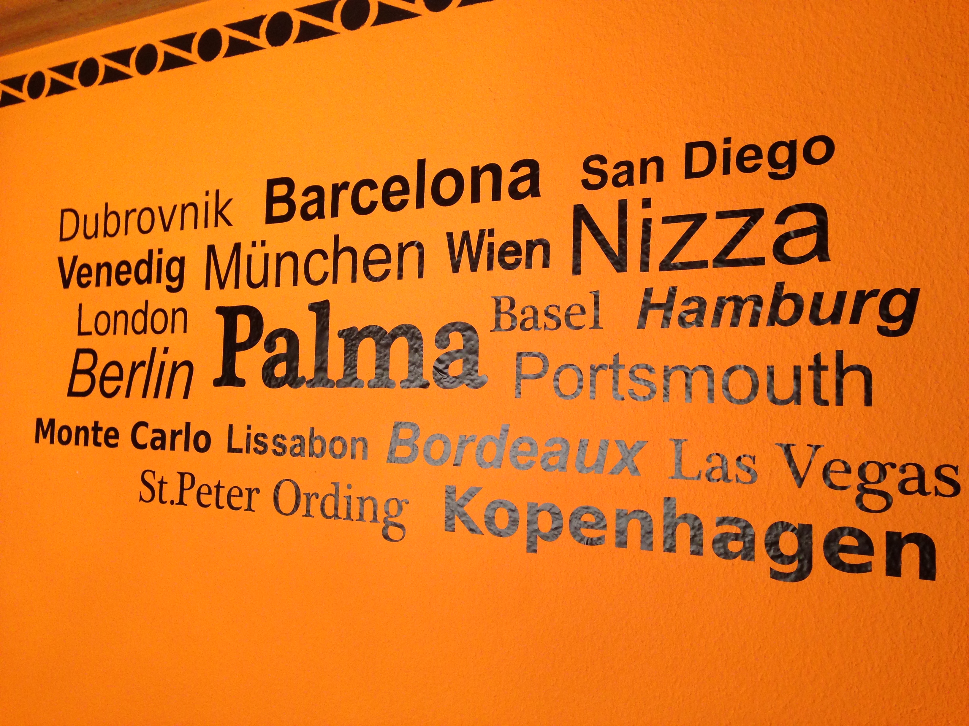 Fertig angebrachtes schwarzes Wandtattoo mit Städtenamen wie Barcelona, Nizza, Palma und Kopenhagen auf oranger Wand, darüber eine Bordüre