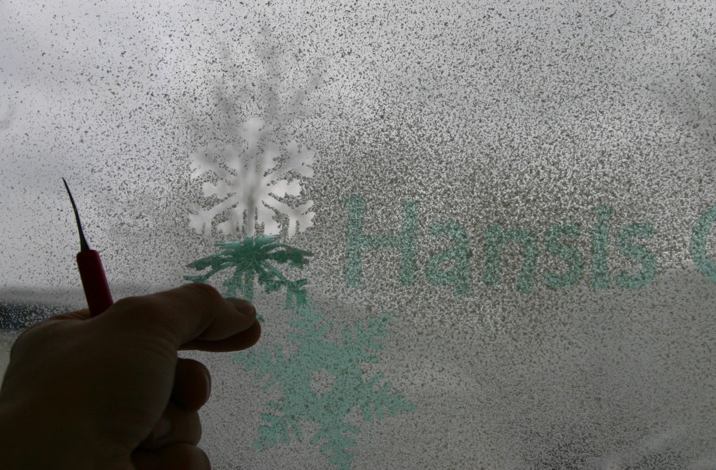Hand löst mit Entgitterhaken grüne Maskierfolie vom mit Snow-Spray besprühten Fenster, darüber eine fertige Schneeflocke