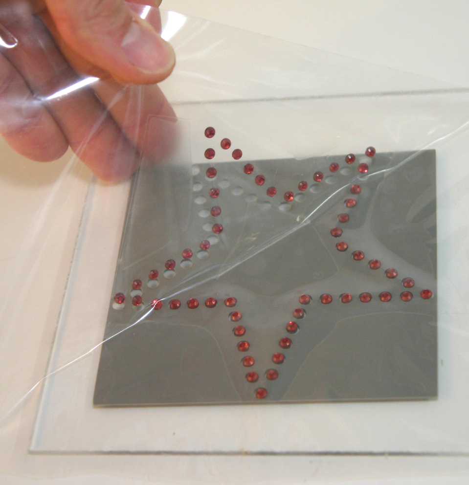 Hand löst Übertragungsfolie von grauer Schablone auf Acrylglas, rote Strass-Steine in Sternform haften an der Folie, leere Löcher bleiben zurück