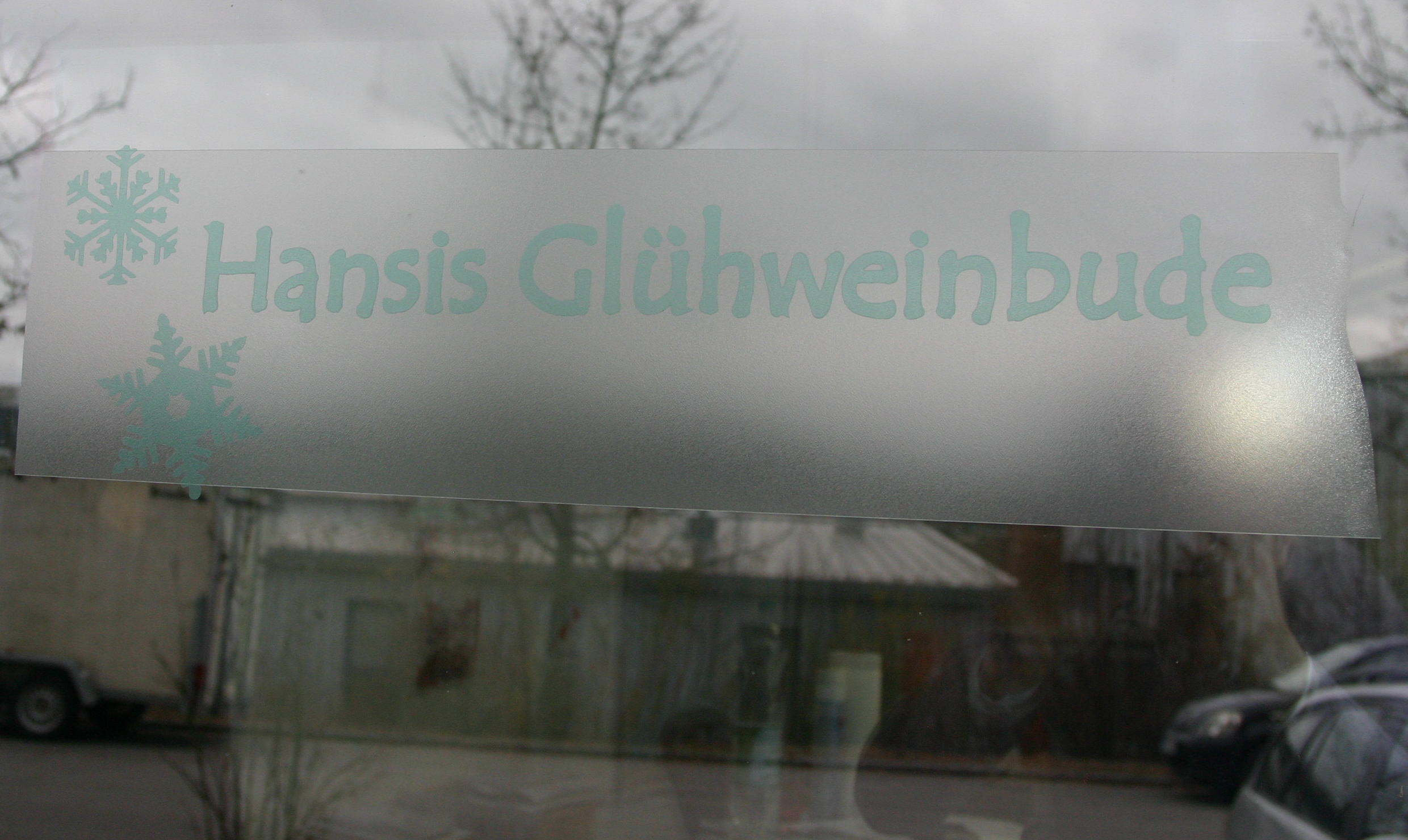 Fensterscheibe mit Snow-Spray besprüht, darauf noch die geplottete Maskierfolie: Schriftzug Hansis Glühweinbude und zwei Schneeflocken