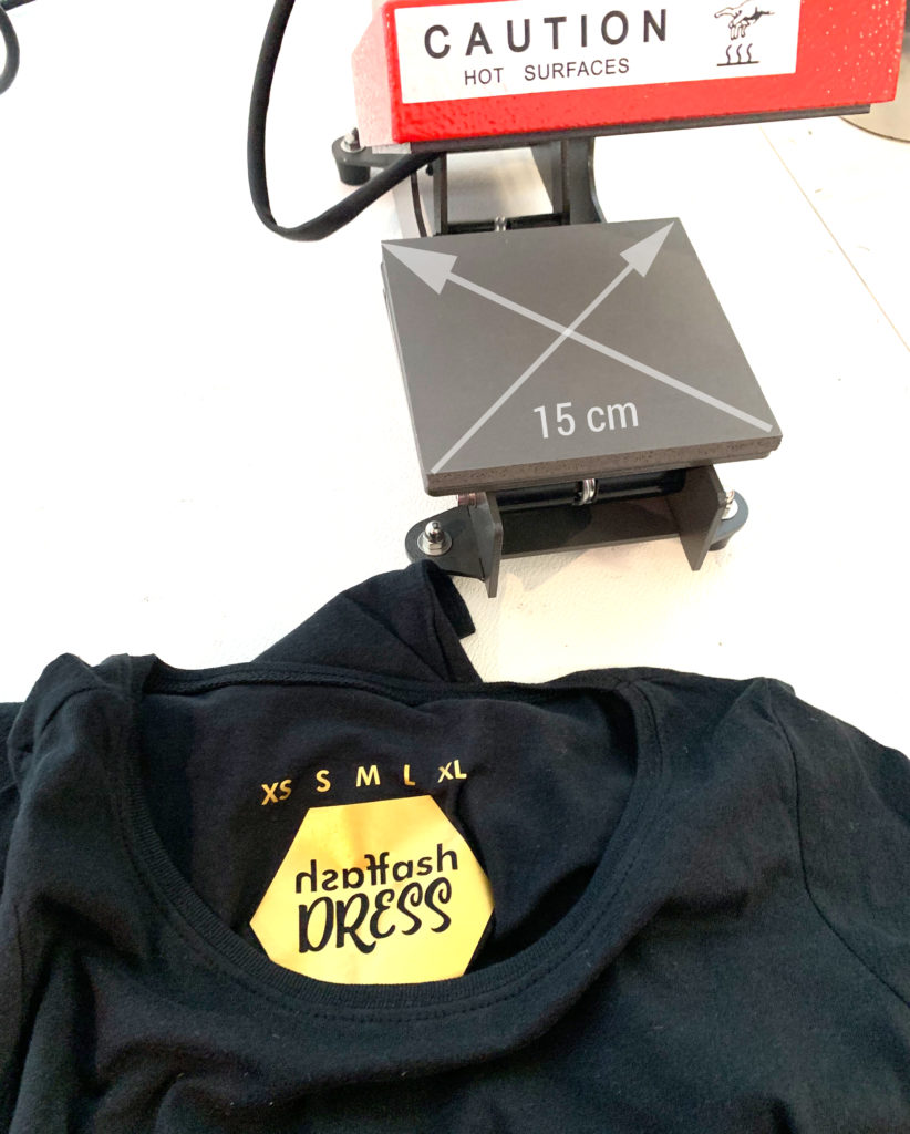 Geöffnete Secabo TC1 mit 15 cm großer Arbeitsfläche, davor ein schwarzes T-Shirt mit goldenem Nackenetikett-Transfer und Größen XS bis XL