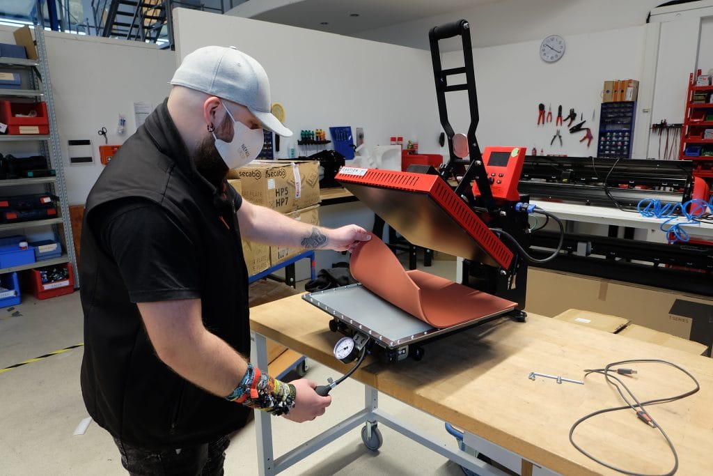 Anwendungstechniker Ääd mit Maske hebt in der Werkstatt die Silikonmatte einer roten Transferpresse an und hält das Manometer
