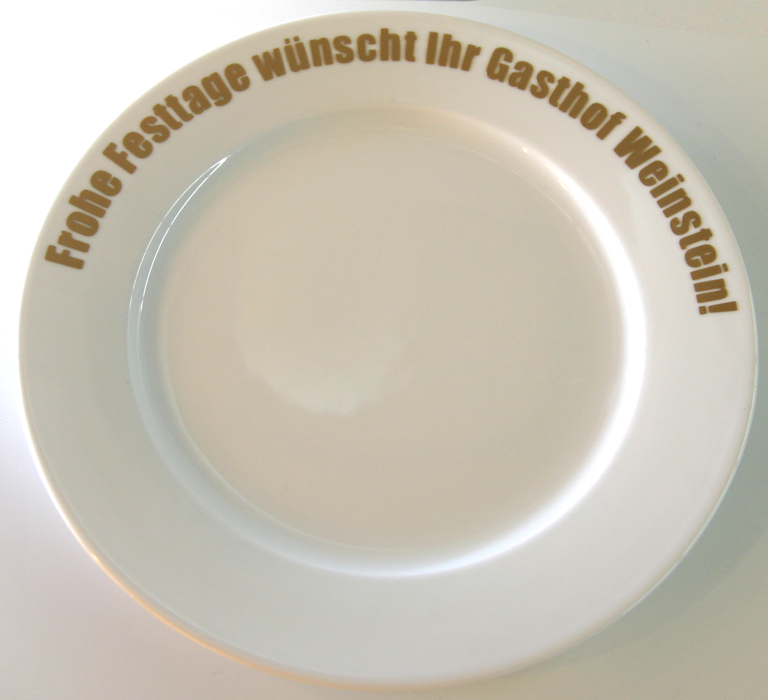 Weißer Teller, auf dem Rand im Bogen ein Schriftzug aus goldener Klebefolie: Frohe Festtage wünscht Ihr Gasthof Weinstein!