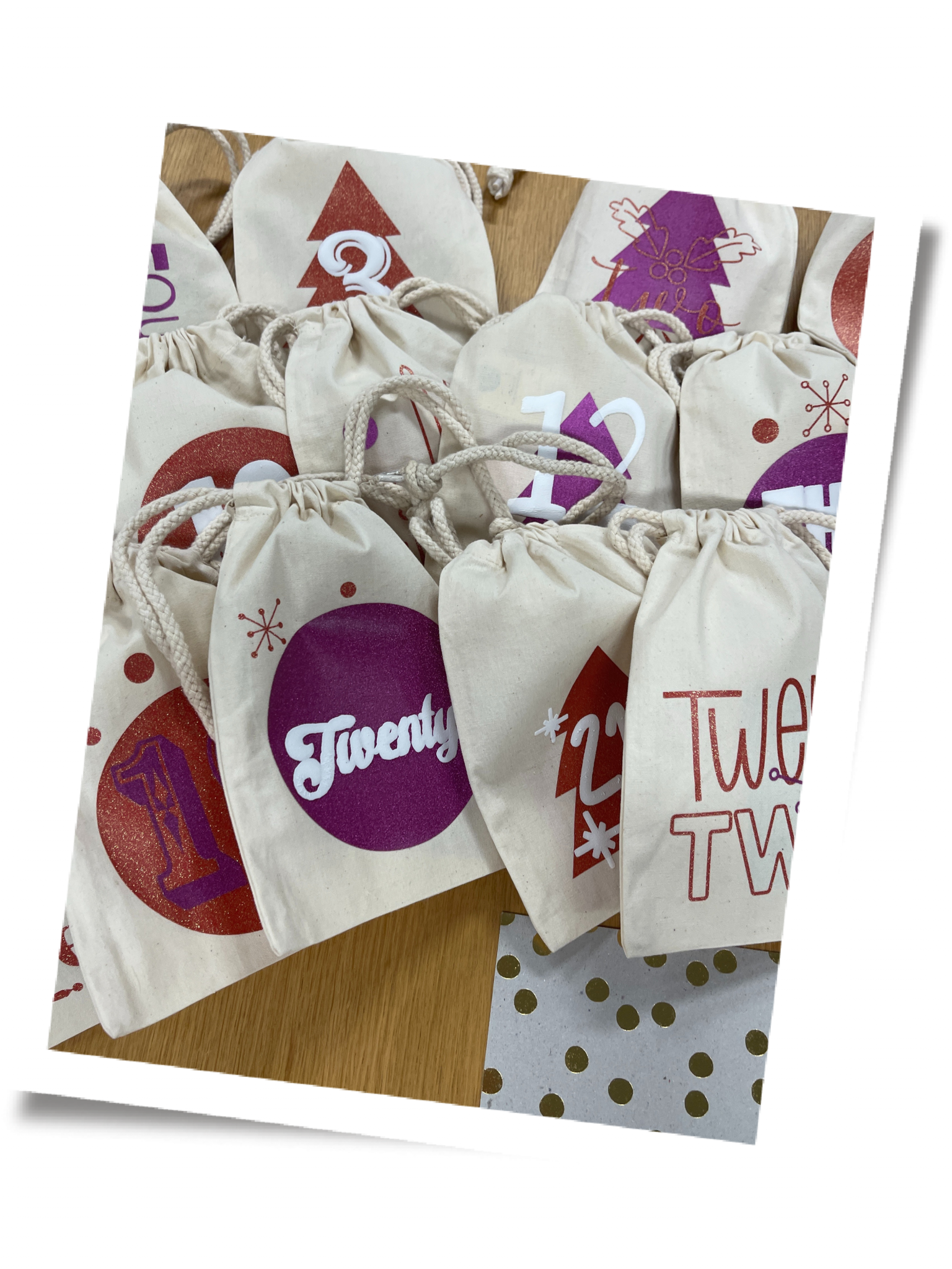 Fertige Adventskalender-Stoffbeutel mit mehrlagig aufgepressten Glitzerfolien in Rot und Pink und weißen Zahlen wie 3, 12 und 22 auf Holztisch
