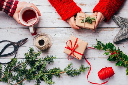 Hände in rotem Pullover halten ein Päckchen in Packpapier, daneben Geschenk mit roter Schleife, Tee, Schnur und Tannenzweige