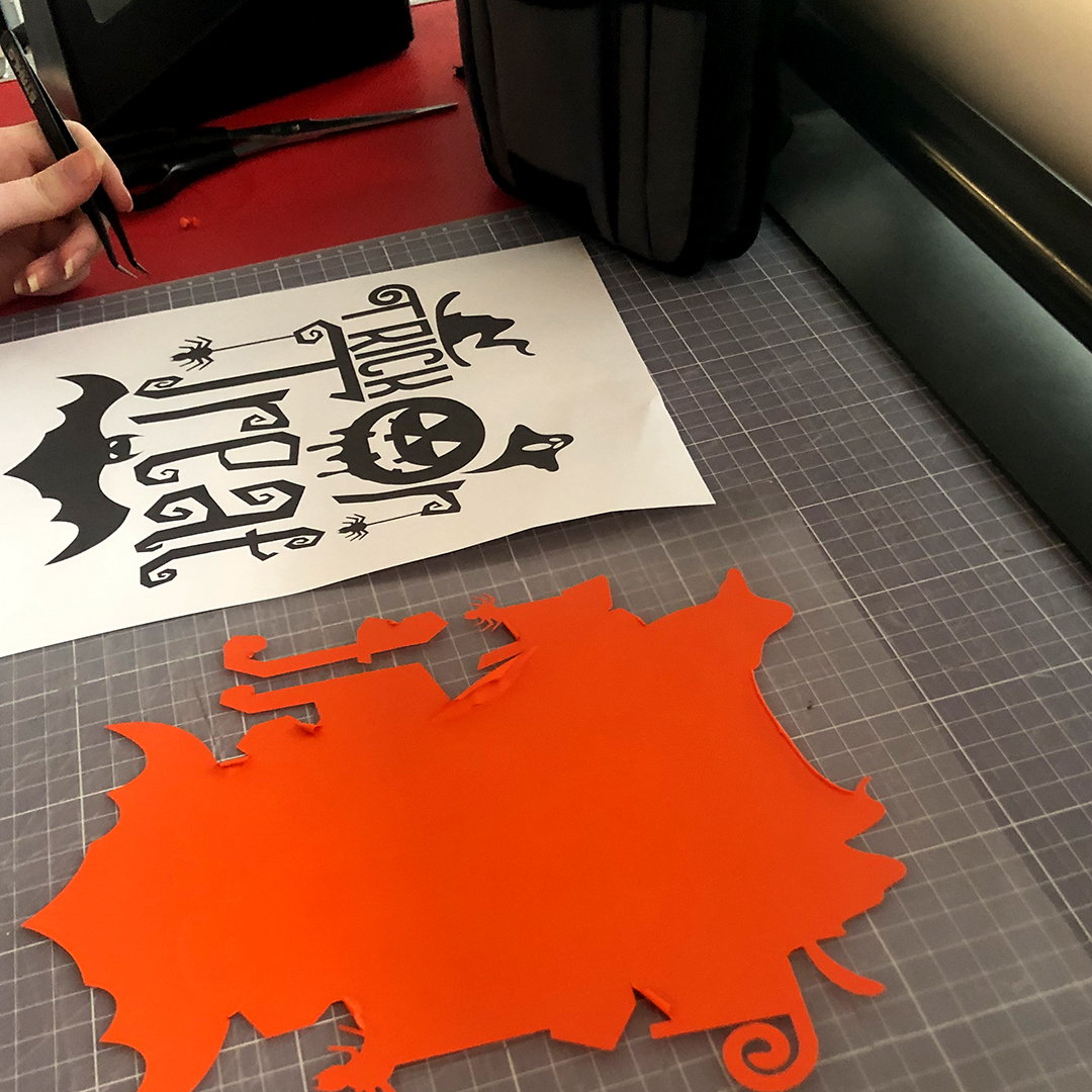 Entgittern: Hand mit Pinzette neben schwarzem Trick-or-Treat-Motiv auf Trägerfolie, davor abgezogene orange Folienreste auf Schneidematte