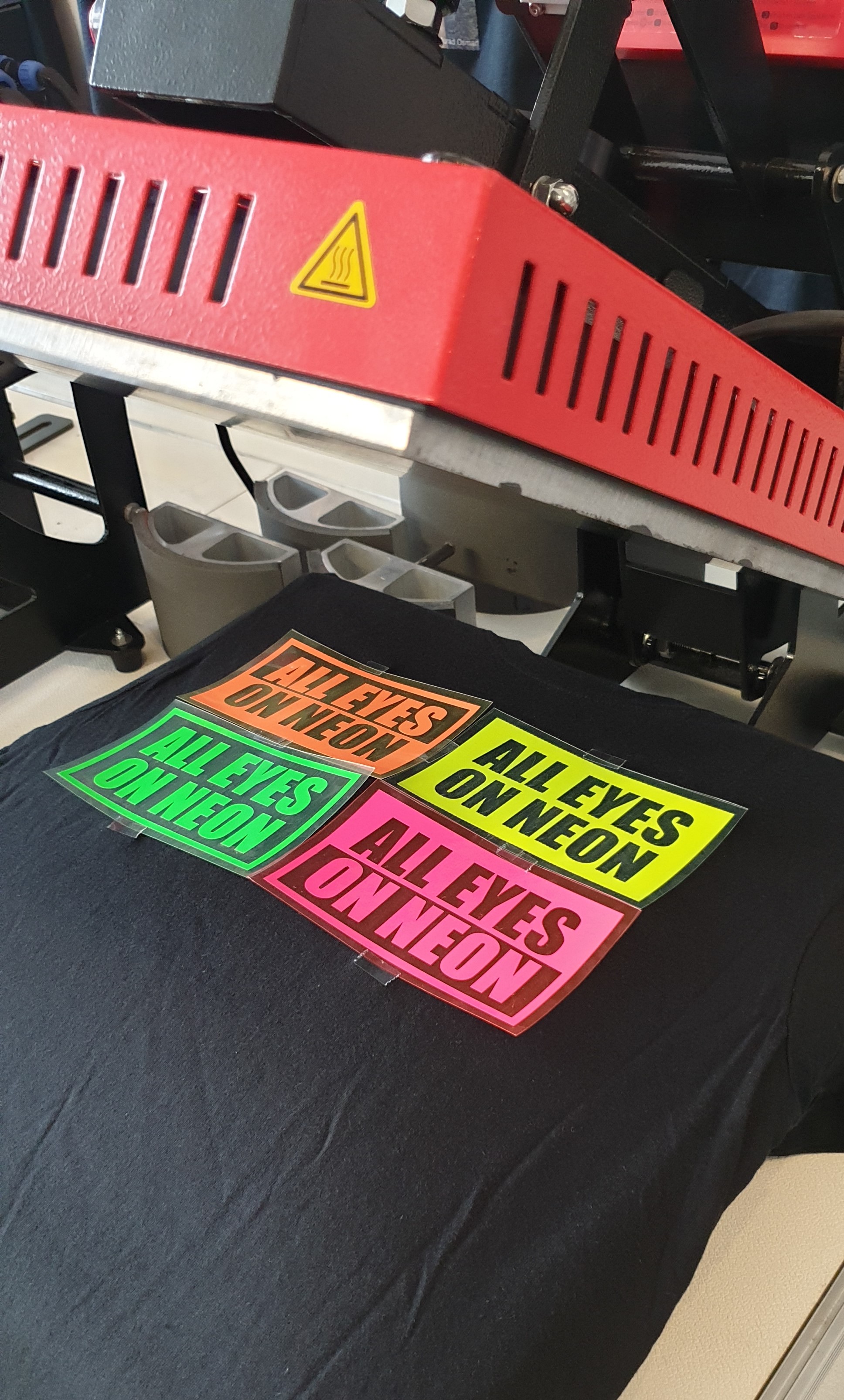 Vier Neon-Flexfolien in Orange, Gelb, Grün und Pink mit Schriftzug ALL EYES ON NEON auf schwarzem T-Shirt unter roter Transferpresse
