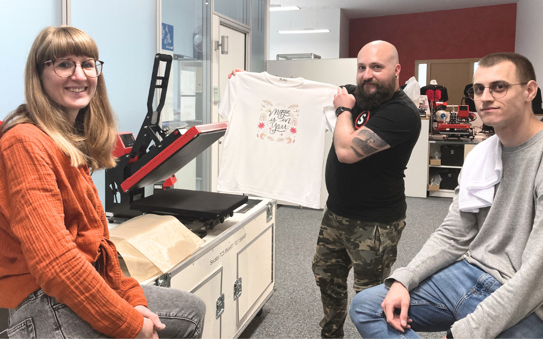 Kathrin mit zwei Kollegen an einer Transferpresse im Büro, ein Kollege hält ein weißes T-Shirt mit dem Aufdruck Magic is in you hoch
