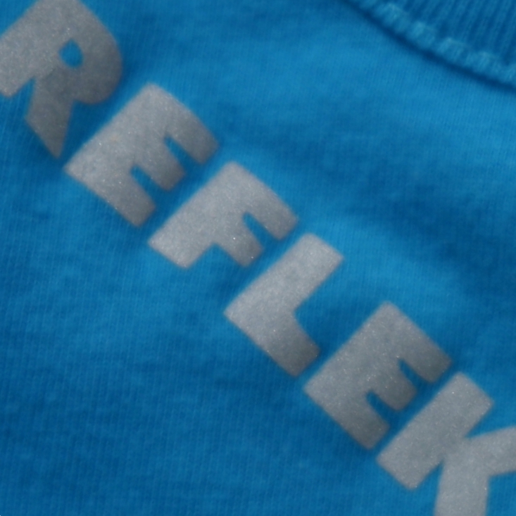 Nahaufnahme: silbergraue reflektierende Flexfolie mit diagonalem Schriftzug REFLEK… auf blaues T-Shirt aufgepresst