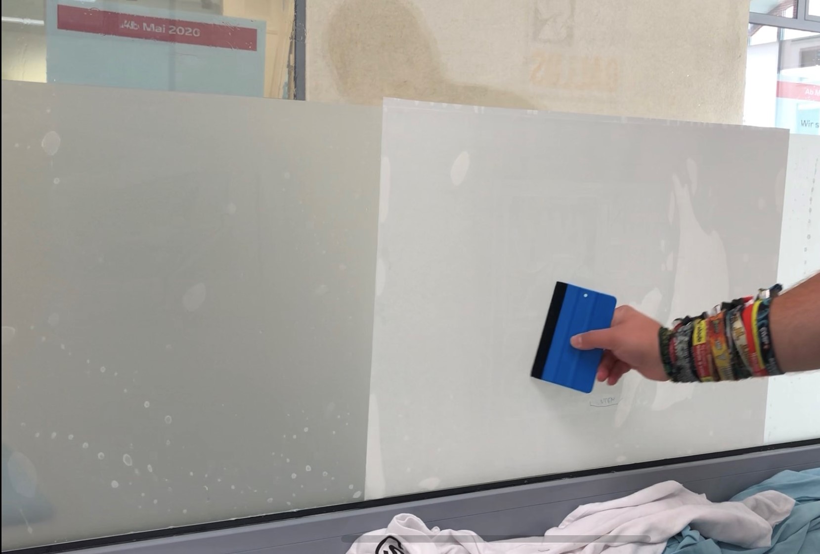 Hand streicht mit blauem Rakel über frisch aufgeklebte, noch feuchte Milchglasfolie am Fenster, um Luftblasen zu entfernen
