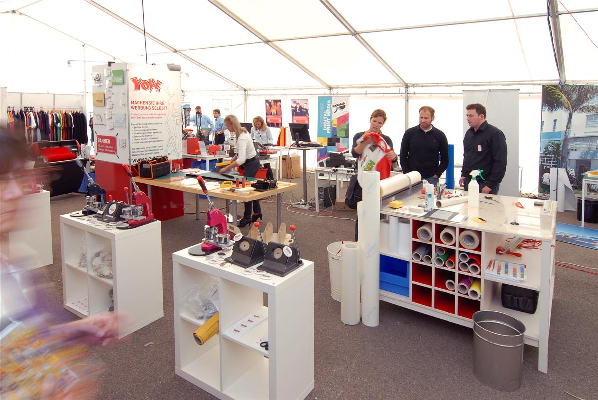 YOW! Hausmesse im Zelt in Wolnzach: Buttonmaschinen, Regal mit Folienrollen und Besucher, die an Stationen beraten werden