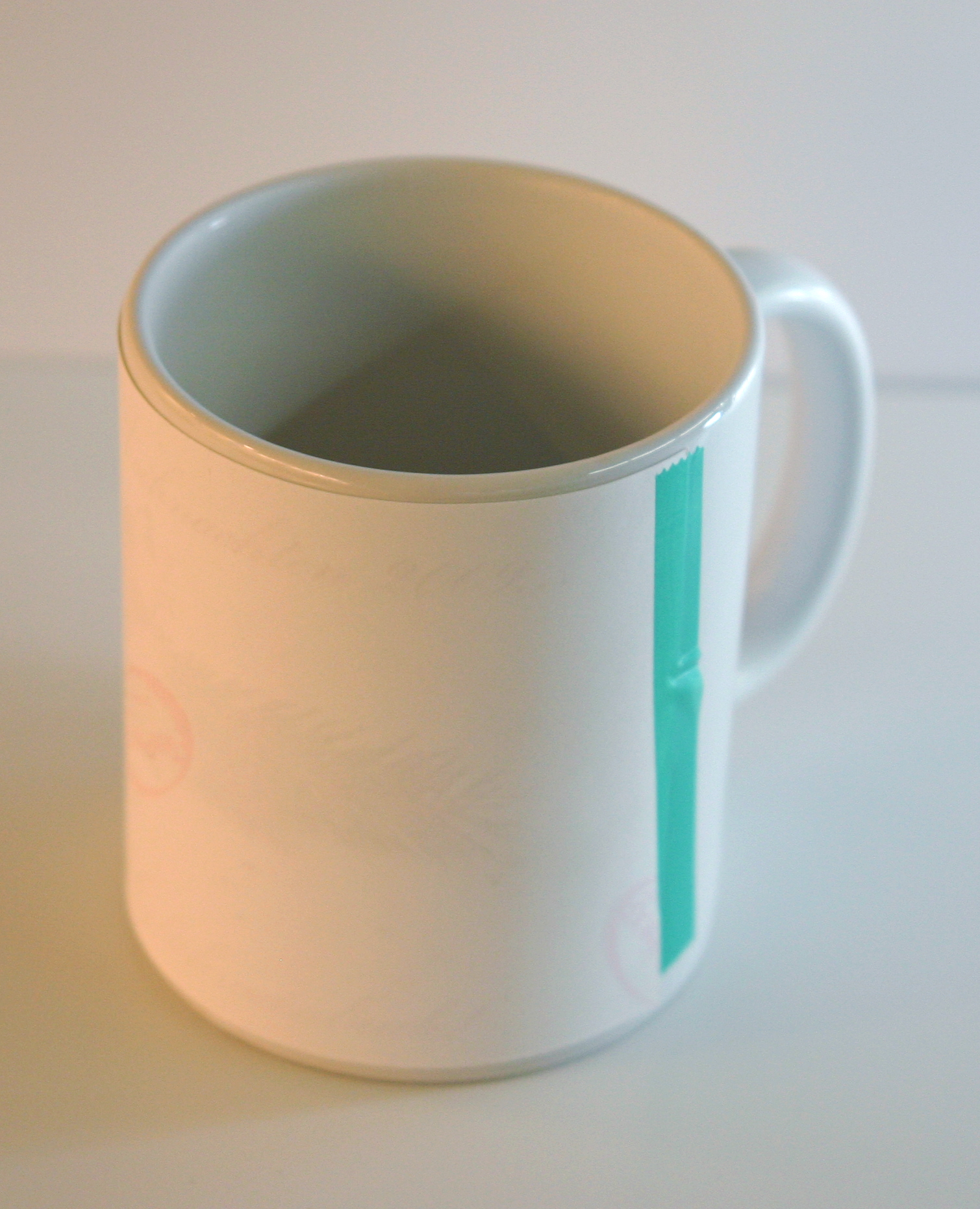 Weiße Tasse, rundum mit bedrucktem Sublimationspapier umwickelt und mit grünem Klebeband fixiert, das Motiv schimmert durch