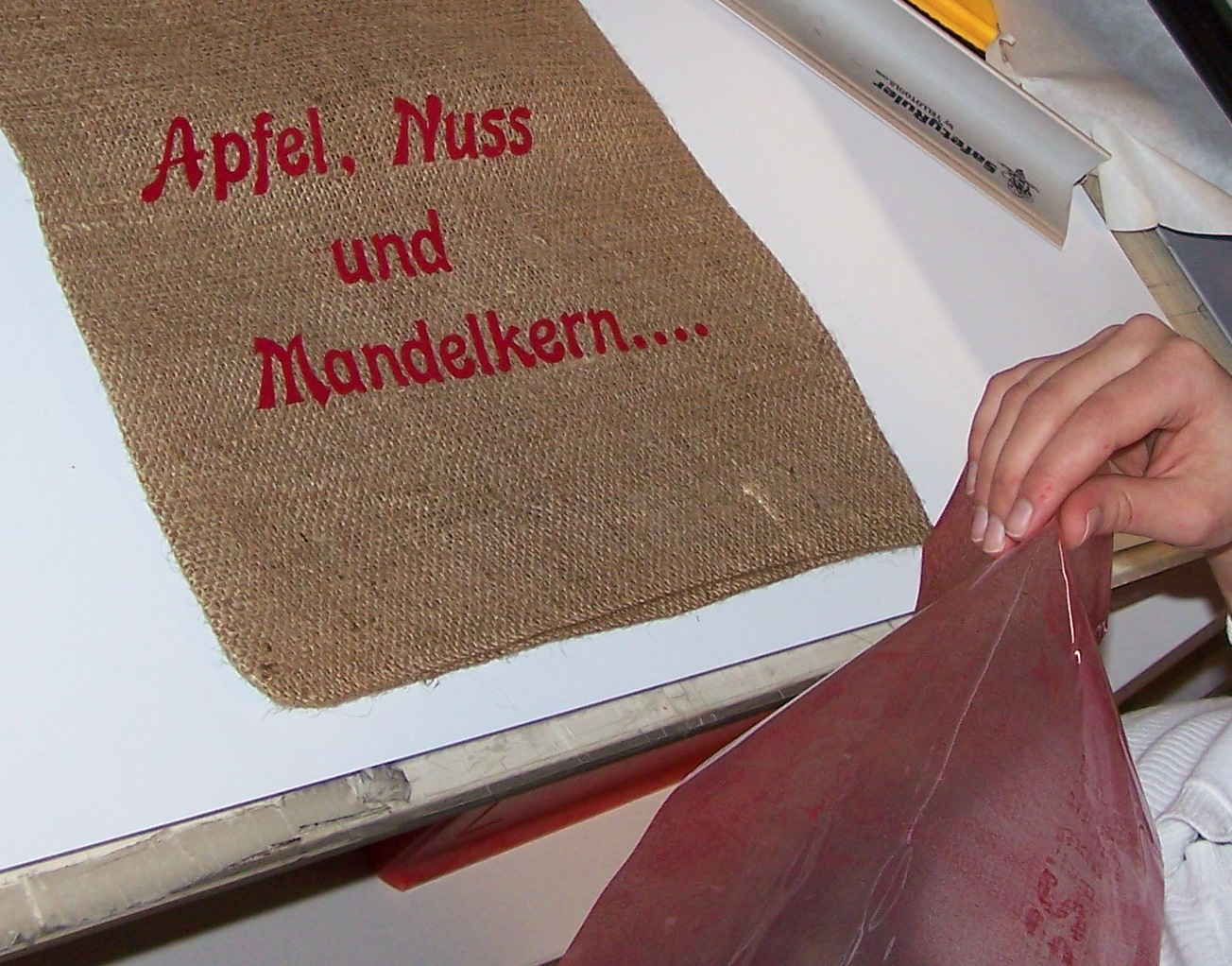 Hand zieht Trägerfolie von einem Jutesäckchen ab, darauf der rote Flockschriftzug Apfel, Nuss und Mandelkern nach dem Aufpressen