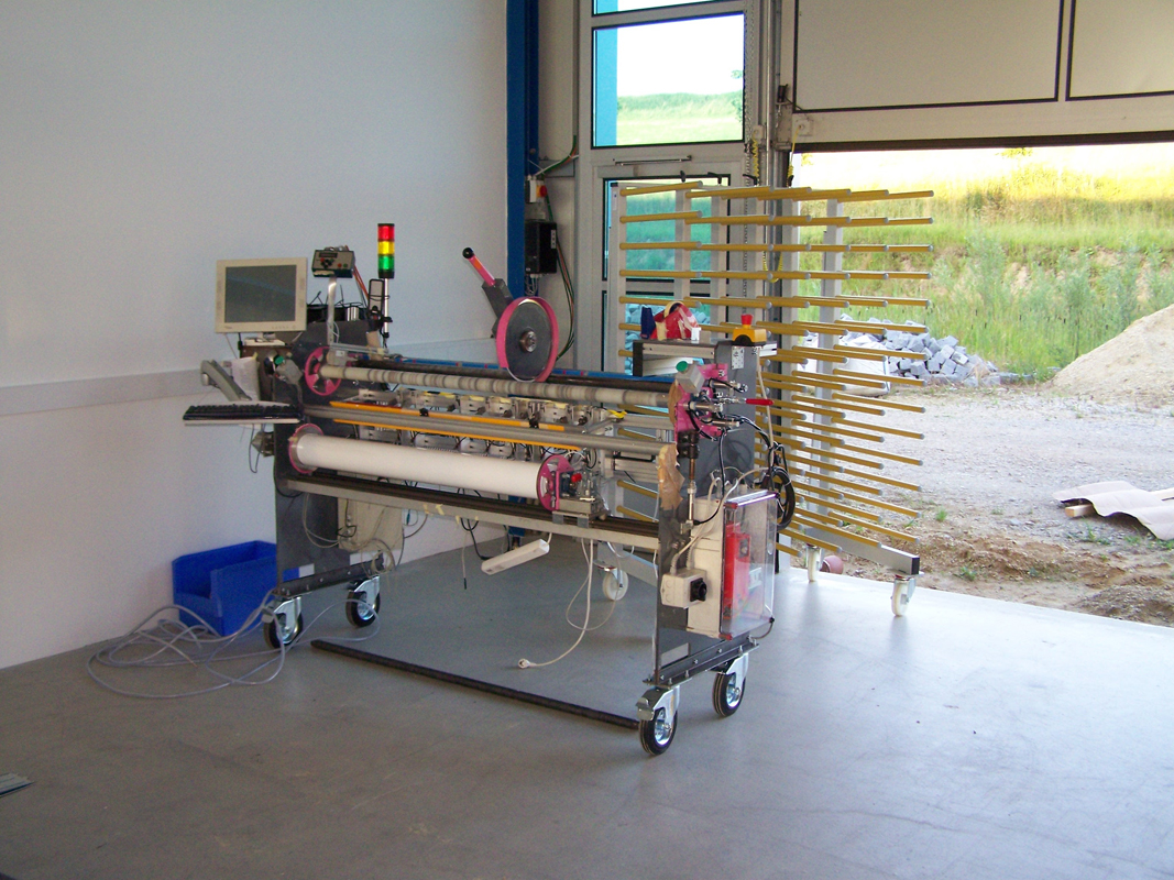 Prototype de la première dérouleuse NEPATA de 2010, montée sur roulettes, équipée d'un écran, d'un voyant lumineux et de rouleaux de films vinyle, dans un hangar ; à côté, un rayonnage à rouleaux
