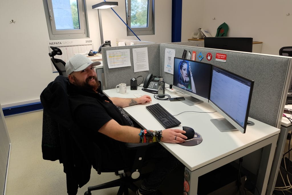 Anwendungstechniker Ääd mit Kappe und Bart sitzt lächelnd an seinem Schreibtisch mit zwei Monitoren im Büro der Nepata Group