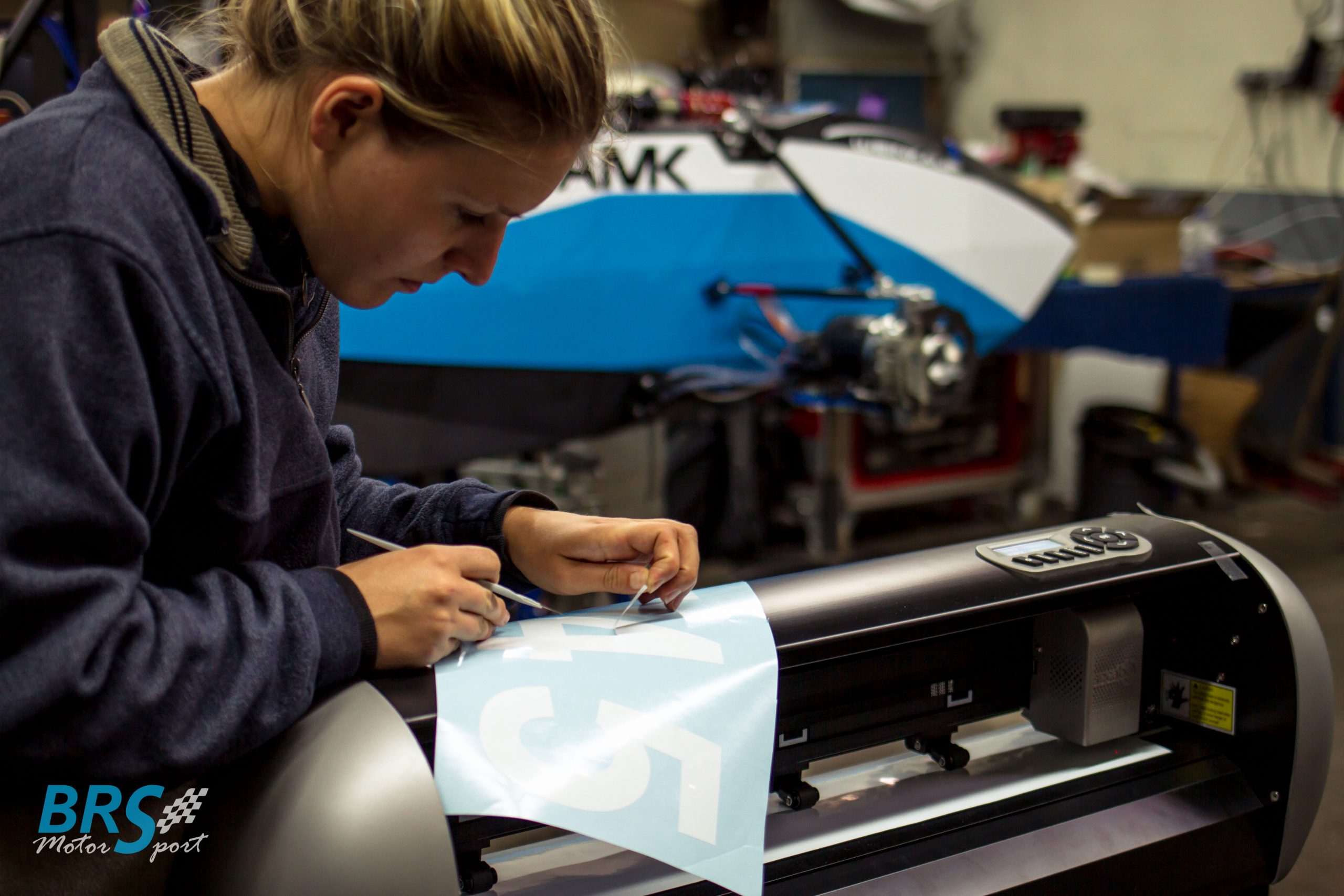 Studentin von BRS Motorsport entgittert eine Startnummer aus weißer Folie am Secabo-Schneideplotter, dahinter der blau-weiße Elektro-Rennwagen