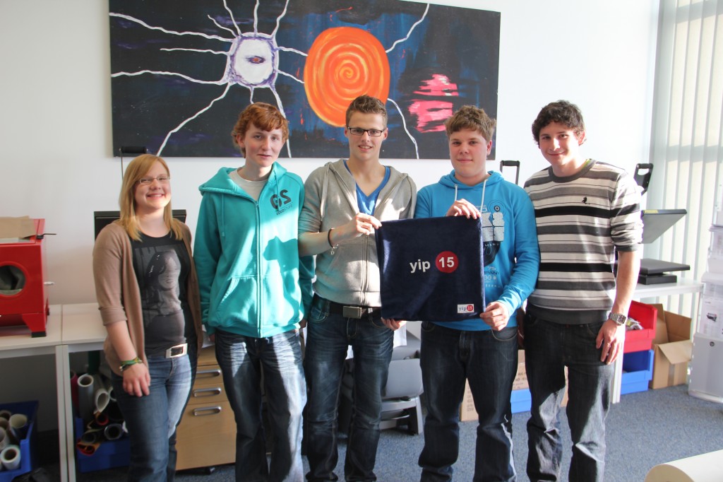 Fünf Jugendliche des Schülerprojekts YIP15 halten ein dunkelblaues Sitzkissen mit yip-15-Logo, im Hintergrund Transferpressen bei YOW!