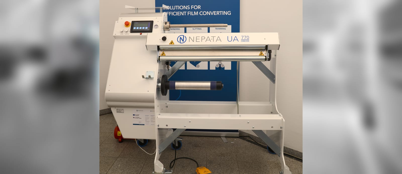 Machine d'enroulement et de découpe NEPATA UA770 Coreless blanche, avec panneau de commande tactile, arbres d'enroulement et pédale de commande, devant une bannière bleue de salon professionnel