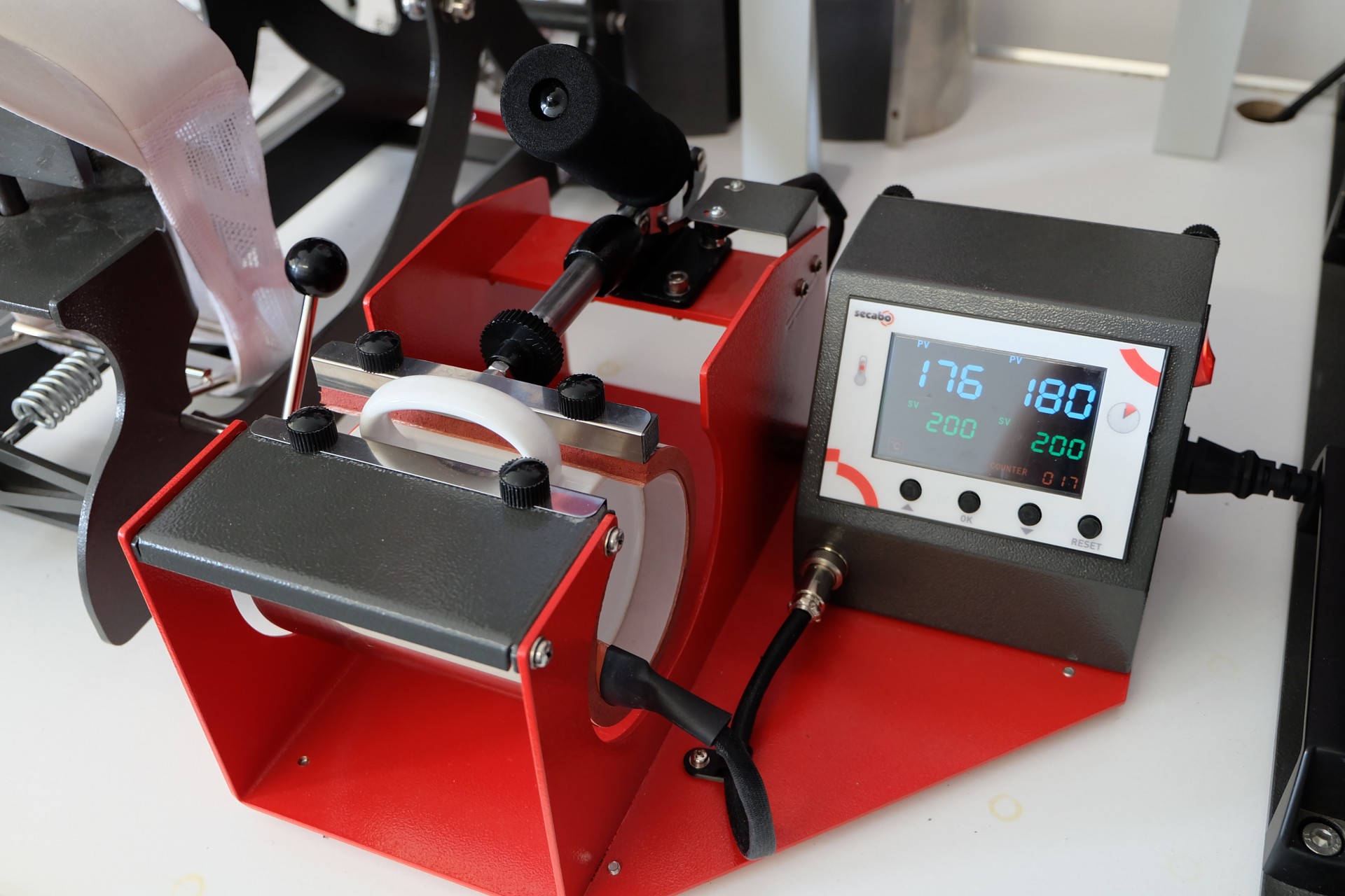 Secabo TM1 Tassenpresse mit eingespannter weißer Tasse, Display zeigt 176 °C bei Soll 200 °C, da die Temperatur beim Pressen zunächst absinkt