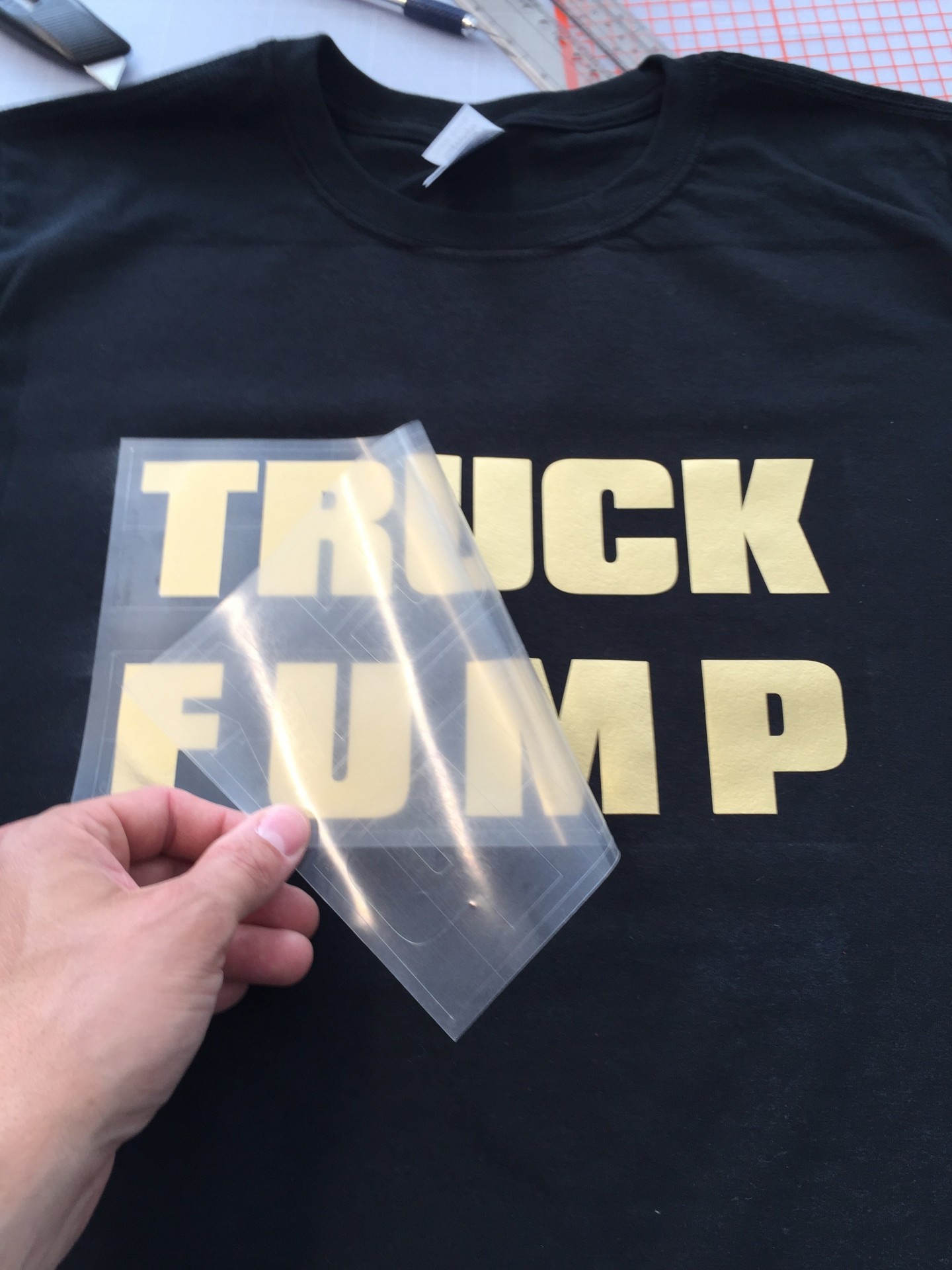 Hand zieht transparente Übertragungsfolie vom goldenen Flexfolien-Schriftzug TRUCK FUMP auf einem schwarzen T-Shirt ab