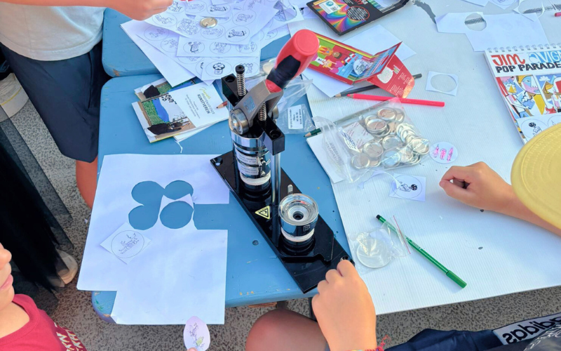 Secabo-Buttonpresse auf einem Basteltisch, umgeben von Kinderhänden, Buttonrohlingen, ausgeschnittenen Papierscheiben und Stiften