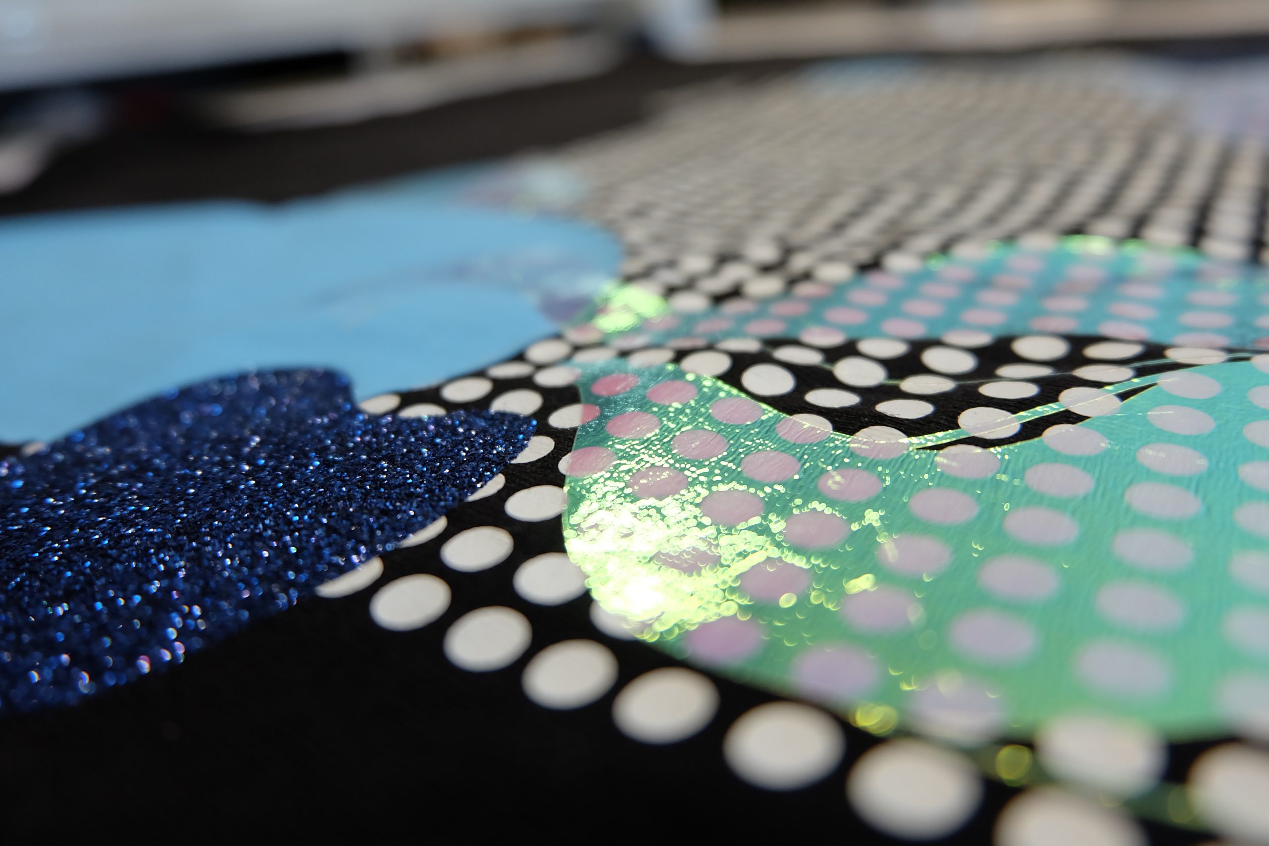 Nahaufnahme eines schwarzen T-Shirts mit aufgepressten Transferfolien: weiße Punkte, blaue Glitterfolie, hellblaue und irisierende grüne Folie
