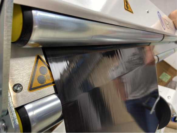 Une feuille d'estampage à chaud brillante défile sans plis et bien tendue sur les rouleaux de la dérouleuse UA1650 ; à côté, un panneau d'avertissement signalant un risque d'entraînement