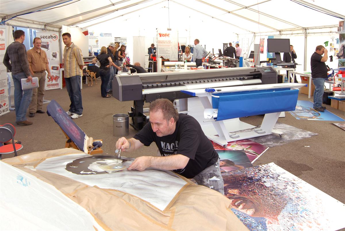 Airbrush-Vorführung auf der YOW!-Hausmesse 2009: Mann sprüht Porträt per Schablone, dahinter Großformatdrucker und Besucher im Messezelt