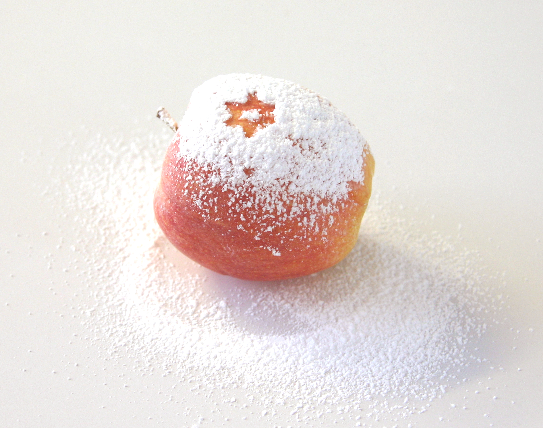 Roter Apfel mit Puderzuckerschicht, in der ein ausgesparter Stern die Schale zeigt, auf weißem Untergrund mit verstreutem Puderzucker