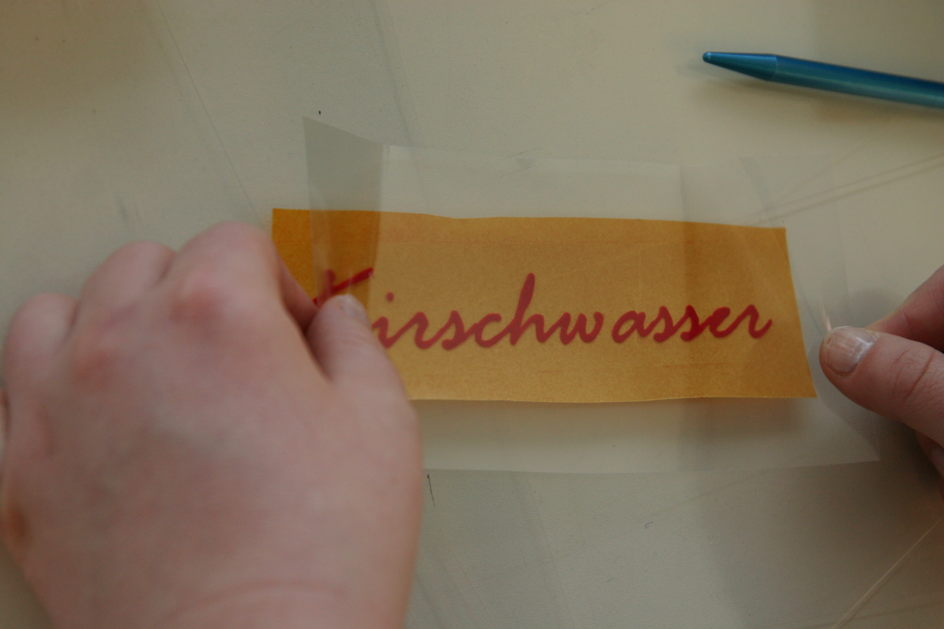 Hände lösen Übertragungsfolie mit rotem Flockfolien-Schriftzug Kirschwasser vom Trägerpapier, um ihn auf ein Objekt zu übertragen
