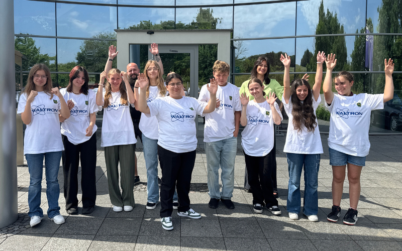 Schüler:innen in selbst veredelten weißen T-Shirts mit WALTRON-Logo winken mit Begleitpersonen vor einem Glasgebäude
