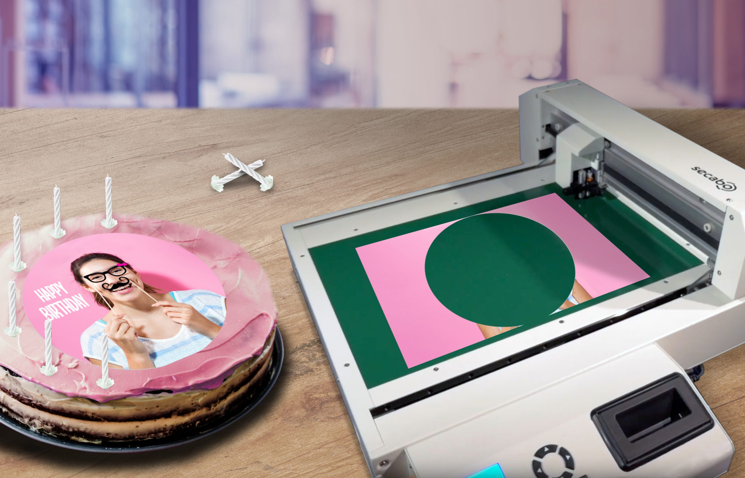 Torte mit rundem Fotoaufleger neben Secabo-Schneideplotter, in dem ein Kreis aus dem bedruckten Esspapierbogen ausgeschnitten ist