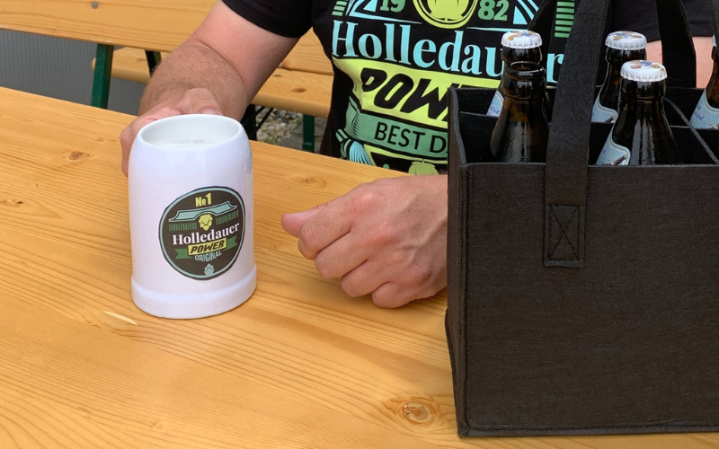 Weißer Keramikkrug mit sublimiertem Holledauer-Power-Logo auf Biertisch, daneben Filz-Flaschenträger mit Bier, Mann im passenden T-Shirt