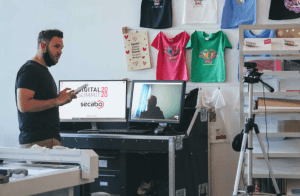 Olly von secabo spricht vor zwei Monitoren mit Digital-Summit-Logo und Videocall, dahinter bedruckte T-Shirts an der Wand