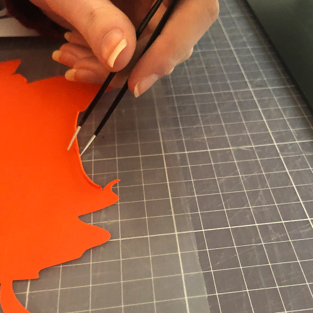 Hand löst mit Pinzette überschüssige orange Transferfolie von der Trägerfolie auf einer Schneidematte, Entgittern des geplotteten Motivs