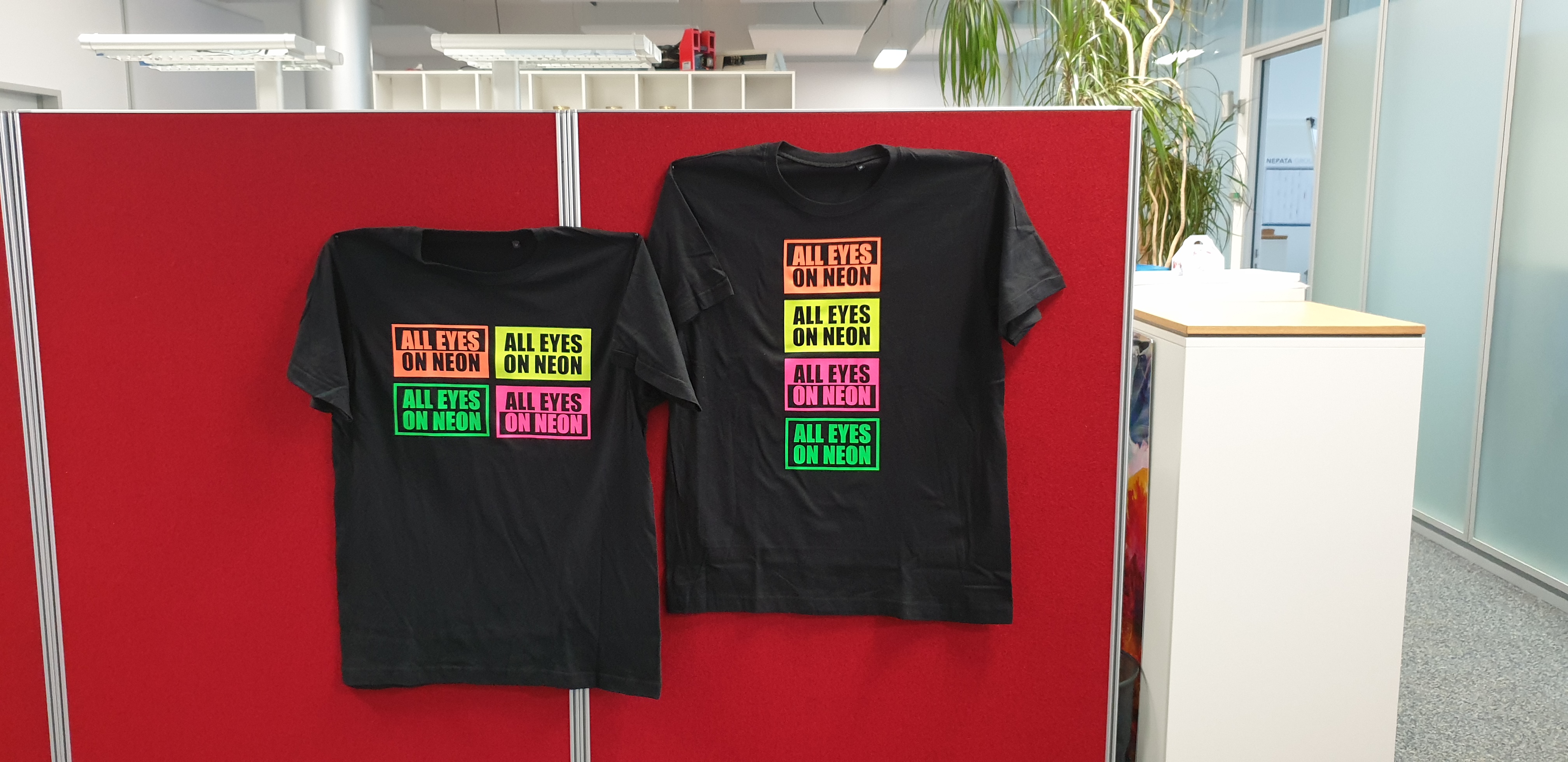Zwei schwarze T-Shirts mit Aufdruck All Eyes on Neon aus Neon Folie in Orange, Gelb, Pink und Grün, an roter Stellwand