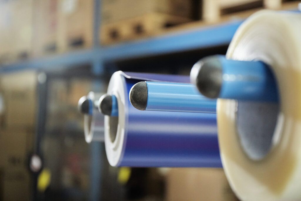 Des rouleaux de films vinyle transparent et bleu sur des mandrins bleus d'un chariot de transport de rouleaux ; à l'arrière-plan, flou, un entrepôt rempli de cartons