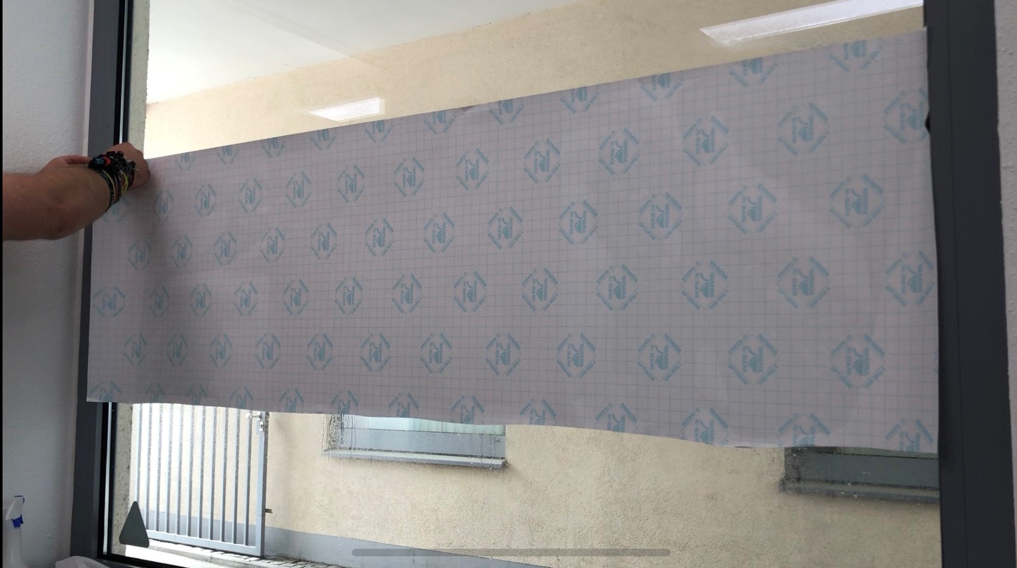 Folie mit Rasterträgerpapier nach außen wird auf befeuchteter Fensterscheibe oberhalb der Zielposition angelegt, eine Hand hält sie fest