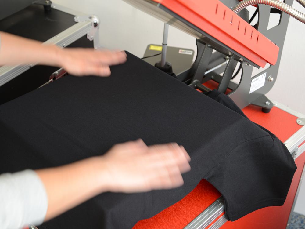 Hände streichen ein schwarzes Baumwoll-T-Shirt auf der Unterplatte einer geöffneten roten Secabo TC5 Transferpresse glatt