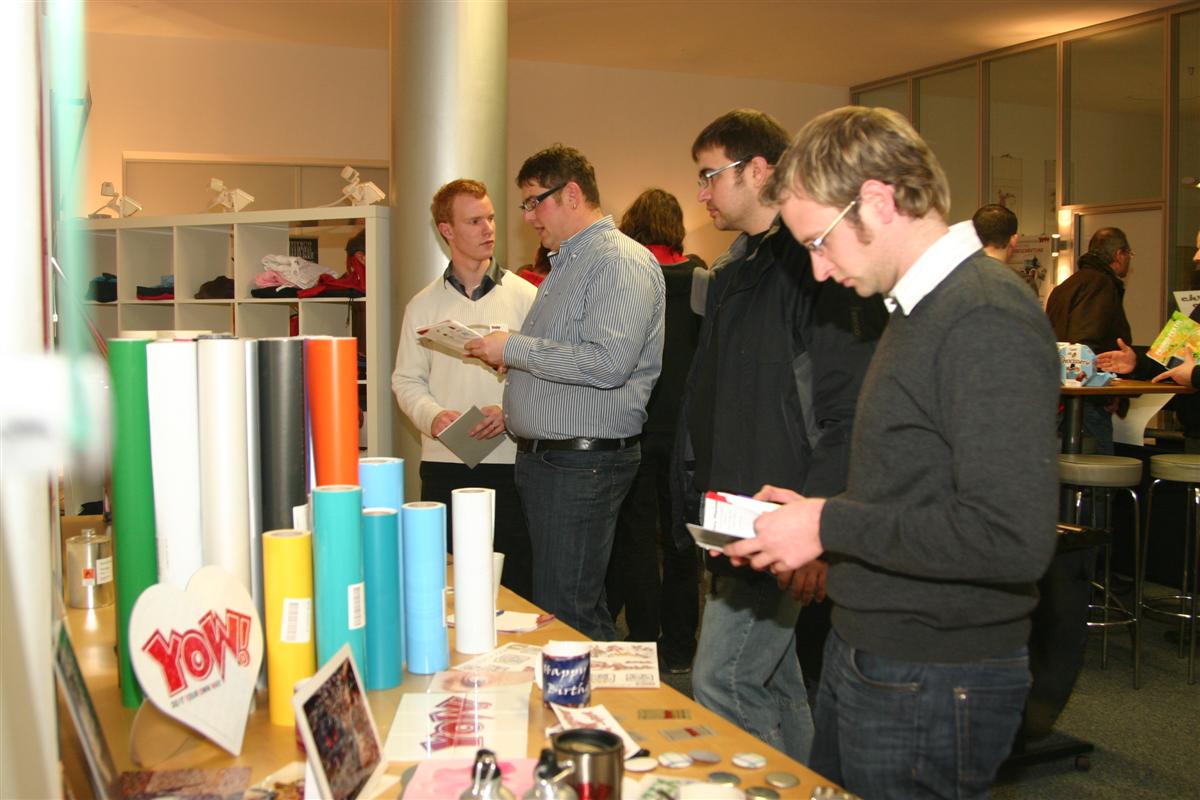 Besucher der YOW!-Hausmesse in Wolnzach lesen Broschüren an einem Tisch mit bunten Folienrollen, bedruckten Tassen und Buttons