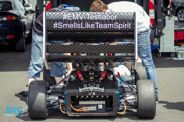 Heckflügel des BRS-Motorsport-Rennwagens mit Folienbeschriftung #EMV-Testlabor und #SmellsLikeTeamSpirit, zwei Teammitglieder am Auto