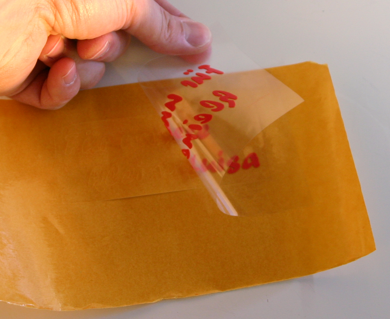 Hand löst Übertragungsfolie mit rotem geplottetem Flock-Schriftzug vom Trägerpapier, bereit zum Aufkleben auf die Wärmflasche