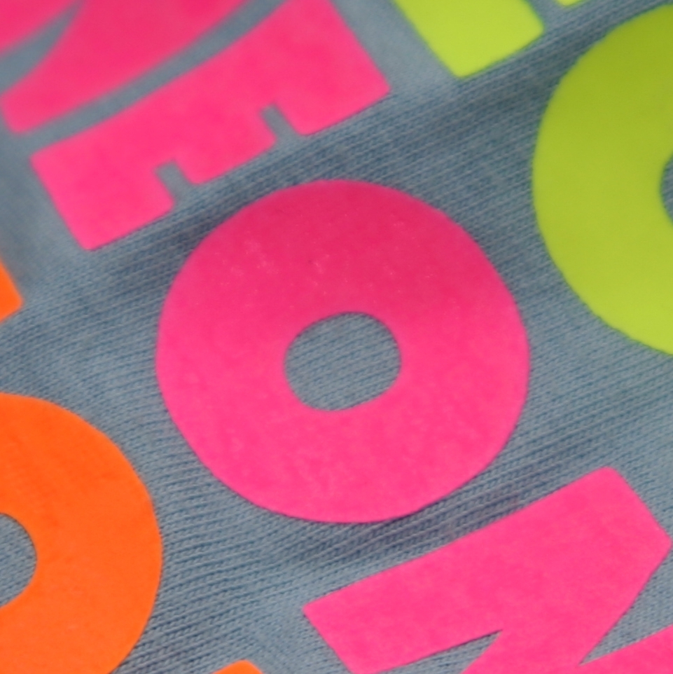 Nahaufnahme aufgepresster Folienbuchstaben in Neonpink, Neonorange und Neongelb auf graublauem Jerseystoff, Oberfläche leicht samtig