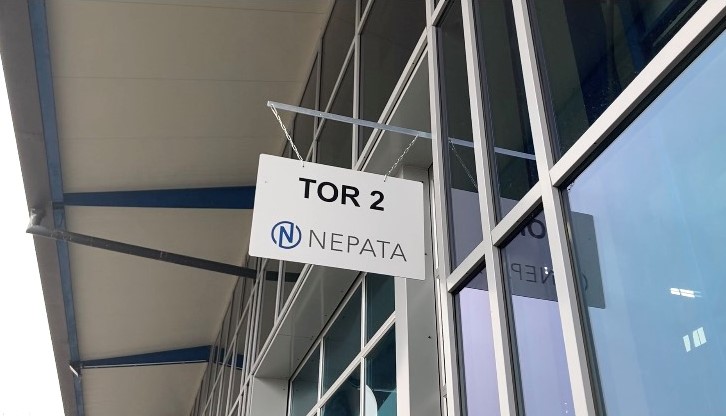 Fertig montiertes weißes Schild mit Aufschrift TOR 2 und NEPATA-Logo, an Ketten unter dem Vordach einer Glasfassade aufgehängt